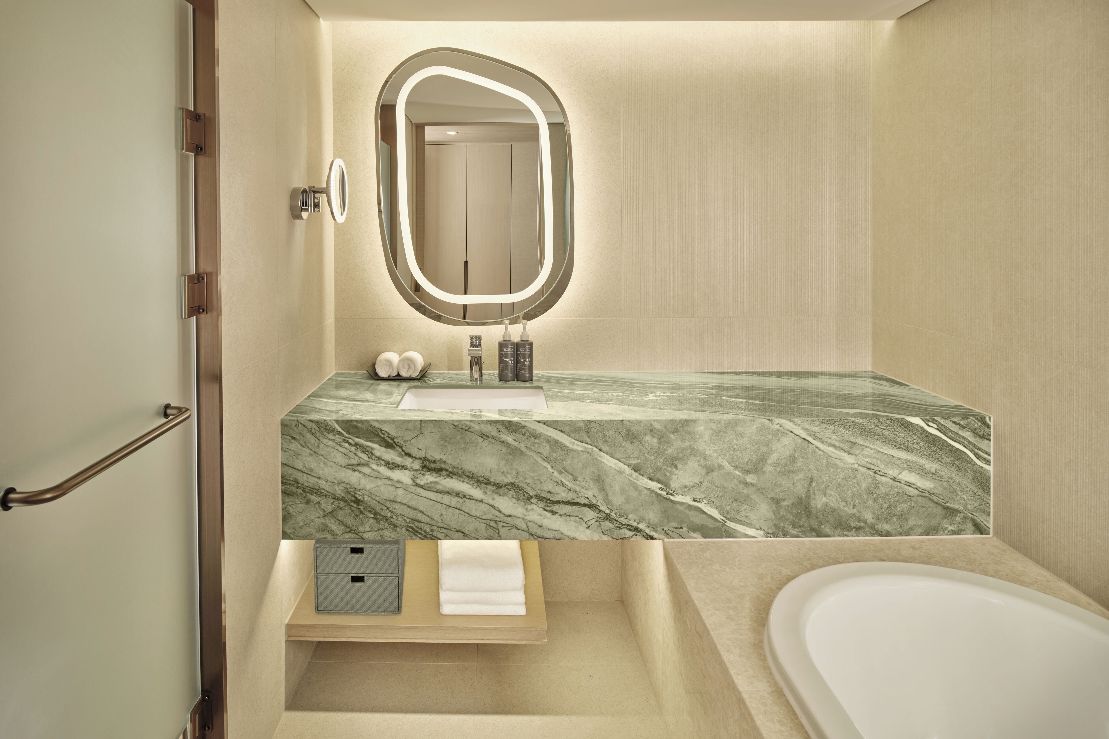 A modern bathroom with a marble sink, a large mirror with lighting, a bathtub, and a towel rack.