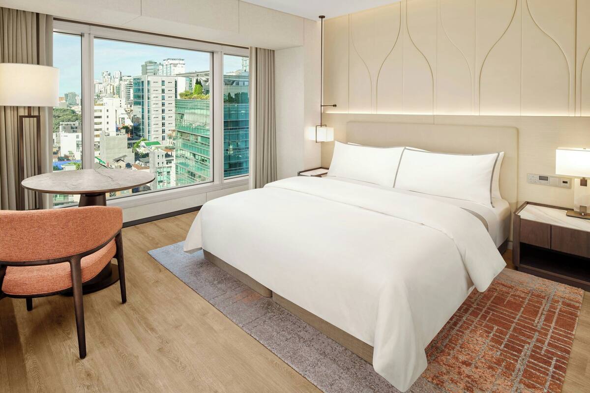 Une chambre d’hôtel avec un grand lit, une petite table, une chaise, une table de chevet et une vue sur la ville grâce à de grandes fenêtres.