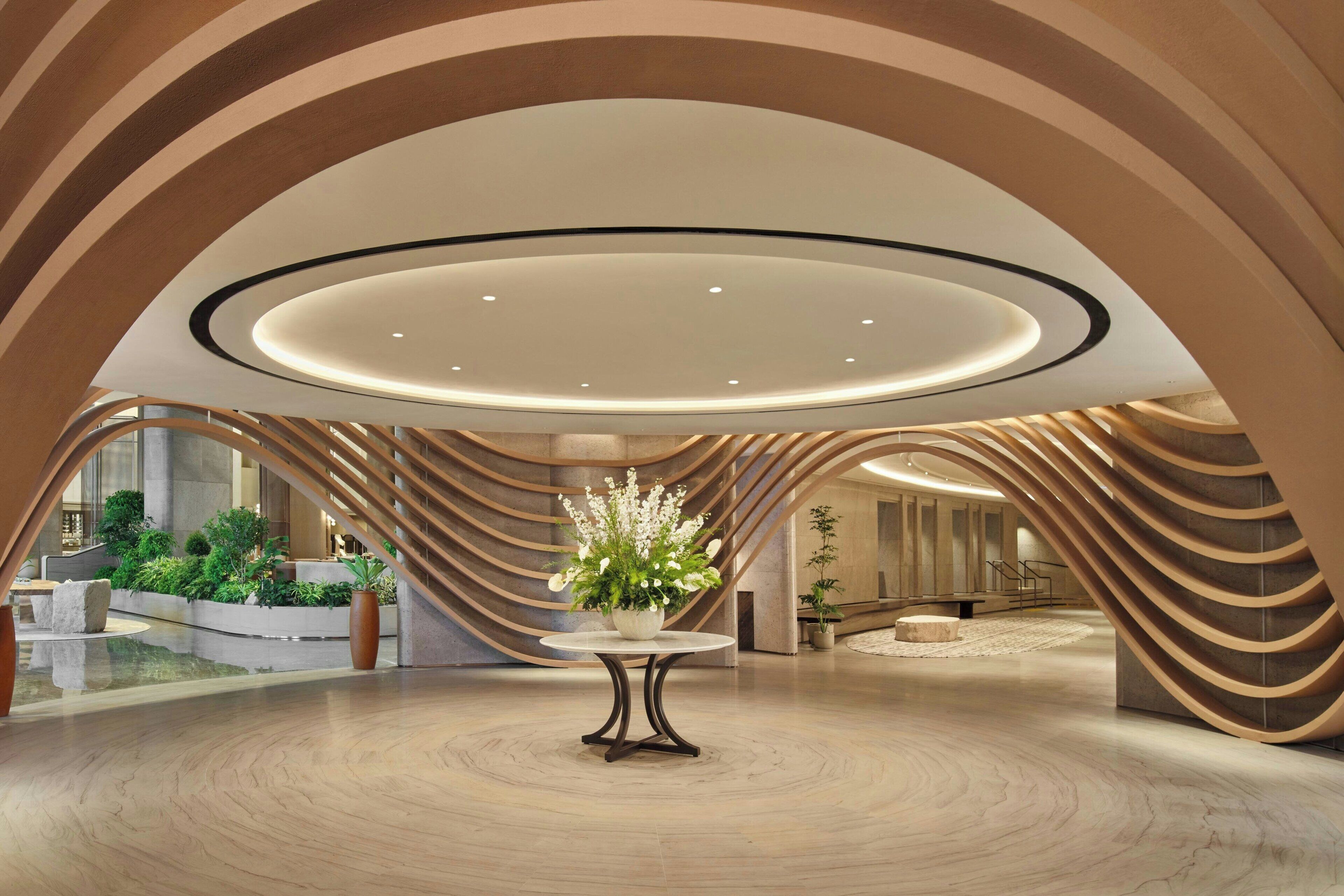 Un hall d’entrée spacieux, doté d’une table ronde centrale, de structures en bois aux lignes courbes et d’un plafond pourvu d’un éclairage encastré.