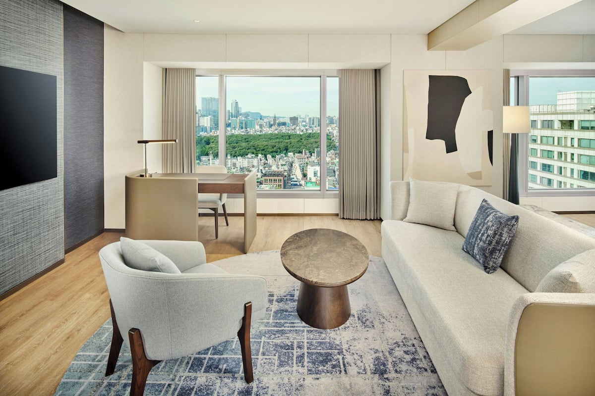 Une chambre d’hôtel moderne dotée d’une grande fenêtre donnant sur la ville, d’un téléviseur à écran plat, d’un canapé, d’un fauteuil et d’une petite table basse ronde.