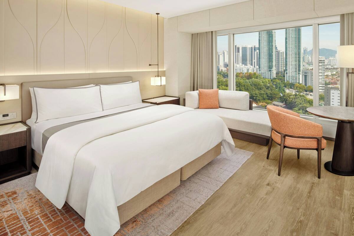 Une chambre d’hôtel avec un grand lit, un canapé, un fauteuil, une petite table et une vue sur la ville grâce à de grandes fenêtres.