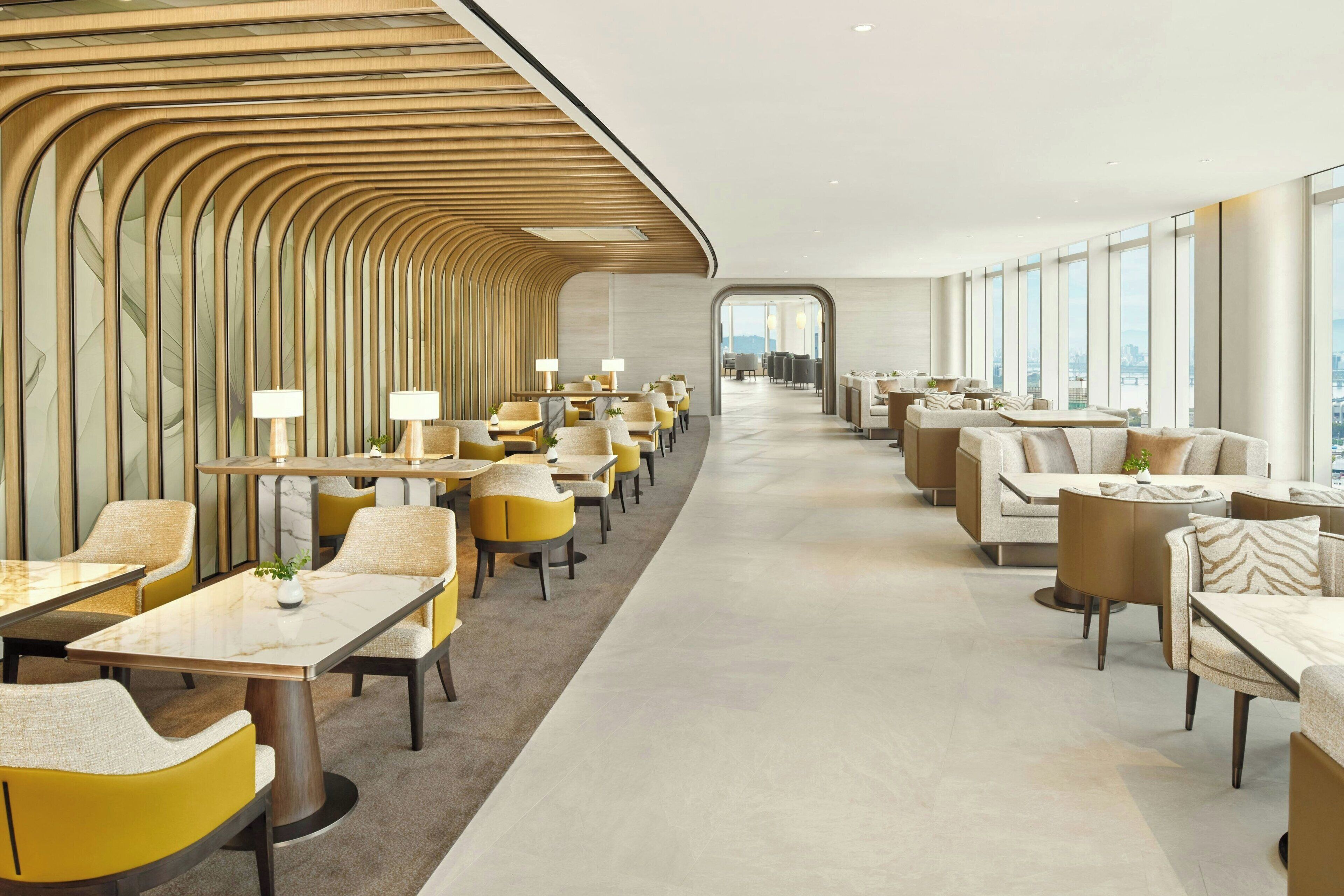 A modern hotel lobby with a long corridor lined with tables and chairs, large windows offering a city view, and a distinctive wooden ceiling design.
