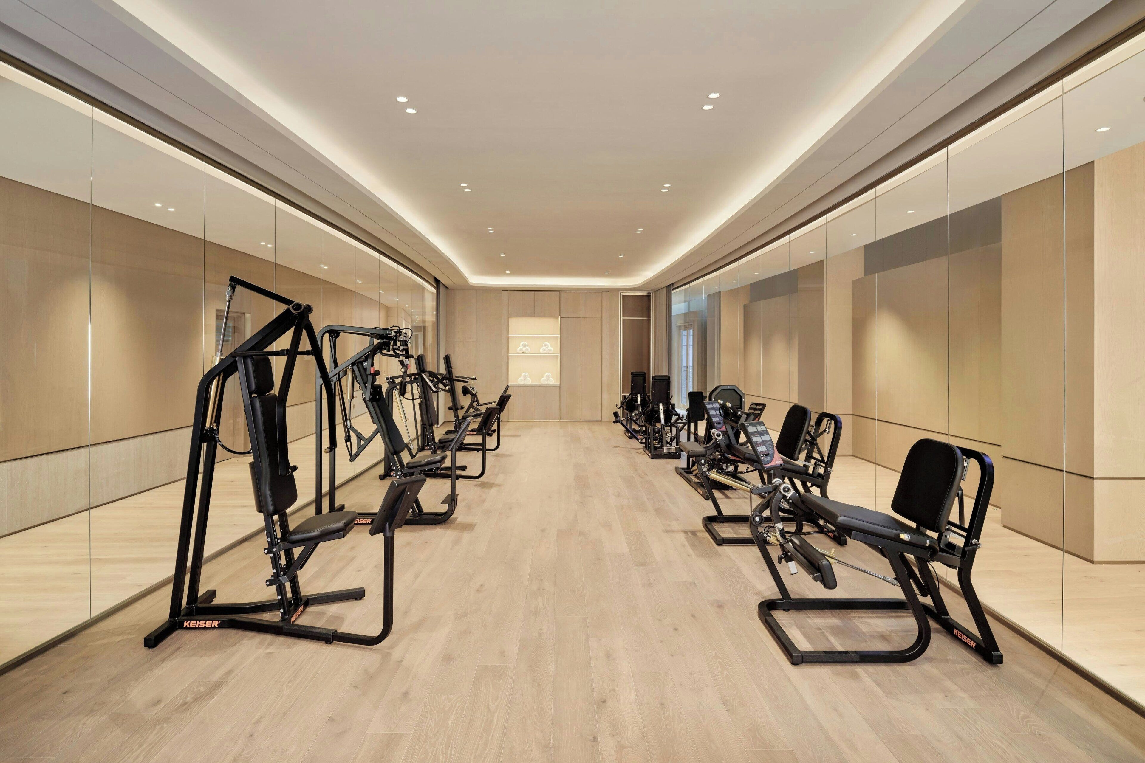 Un gimnasio moderno con diversos equipos de ejercicio, incluyendo cintas de correr y máquinas de pesas, dispuesto en un espacio amplio con suelo de madera y paredes con espejos.