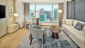 A modern hotel room with a large window, a sofa, a chair, a coffee table, and a TV.
