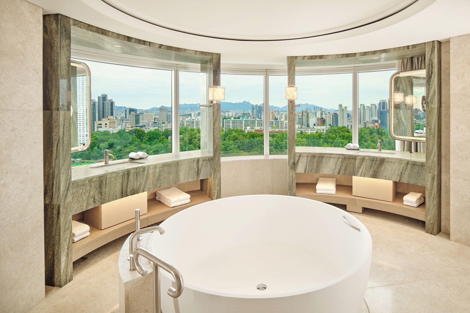 A modern bathroom with a large bathtub, marble walls, and a city view.