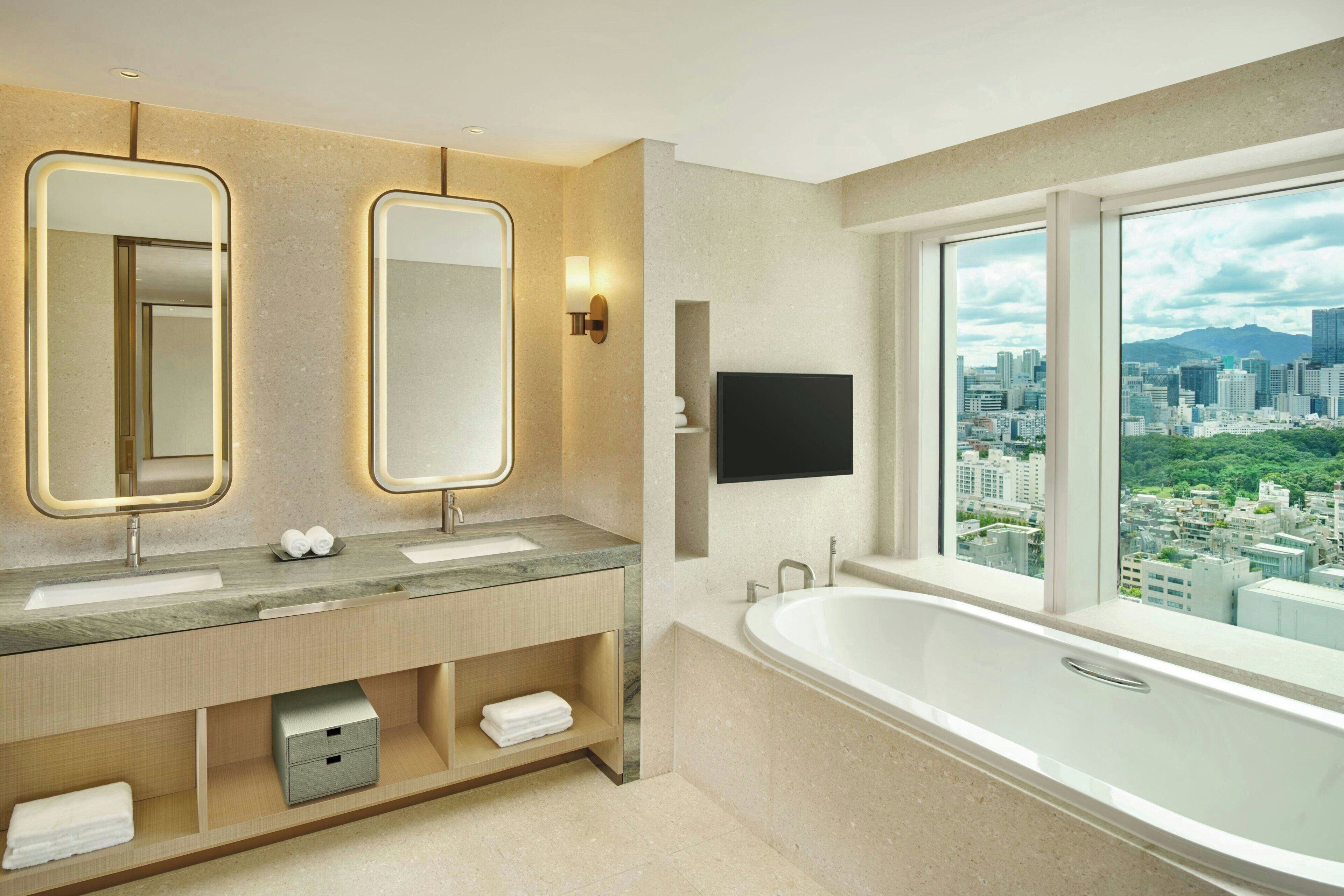 A modern bathroom with dual sinks, a large bathtub, and a city view.