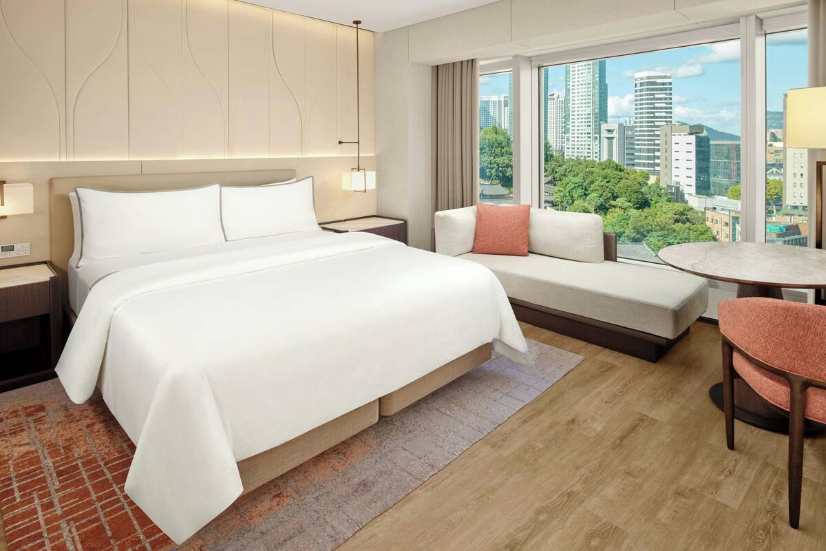 Une chambre d’hôtel avec un grand lit, un canapé, une petite table et une chaise. Il y a une fenêtre donnant sur la ville.