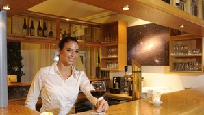Une femme se tient au comptoir d’un bar, avec des verres à vin et des bouteilles en arrière-plan.