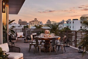 Terraza en la azotea con una mesa de comedor dispuesta para seis personas, sillas alrededor y vistas a un paisaje urbano al atardecer.