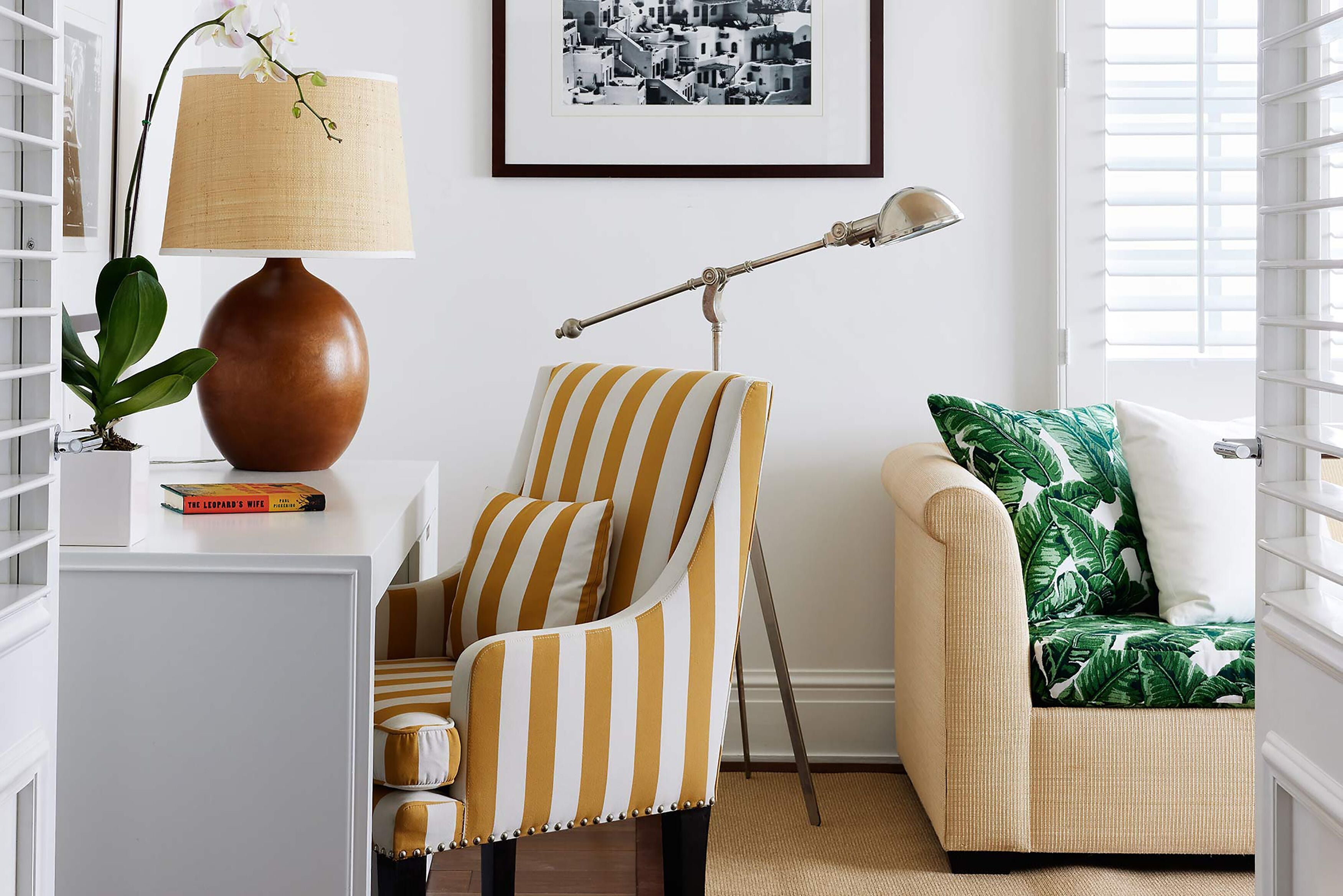 Ein Zimmer mit einem gestreiften Sessel, einem Sofa mit Blätermuster, einer Lampe und einem gerahmten Bild an der Wand.