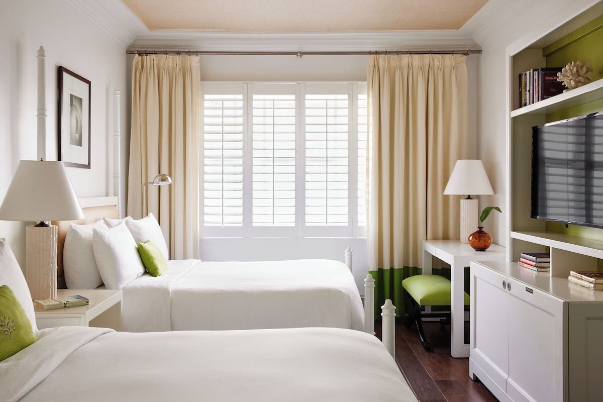 A hotel room with two beds, a desk with a lamp, a television, and a bookshelf.
