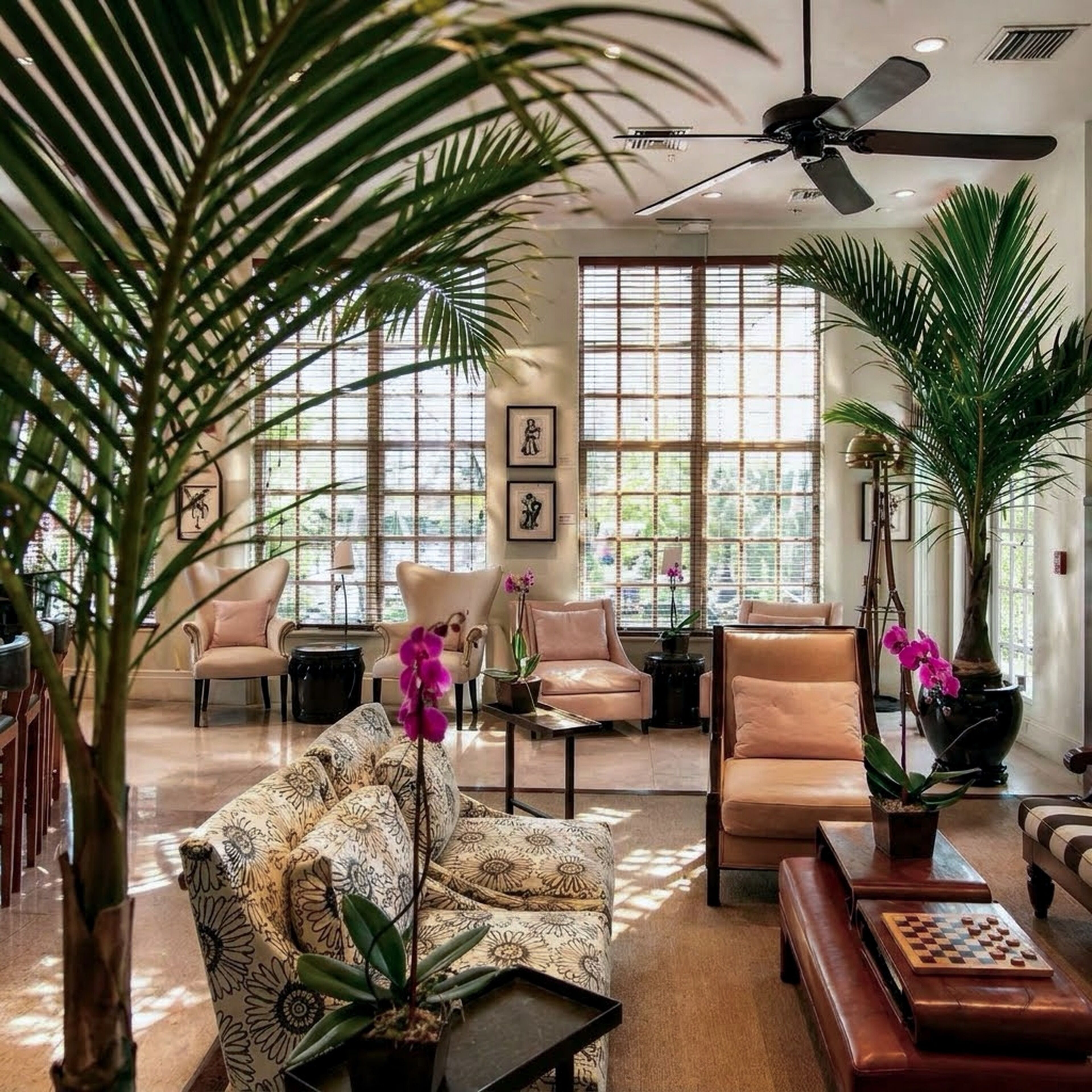 A spacious living room with a ceiling fan, large windows, and various seating arrangements including a patterned sofa and armchairs.