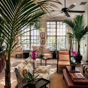Un salon spacieux doté d’un ventilateur de plafond, de grandes fenêtres et de différents types de sièges, notamment un canapé à motifs et des fauteuils.