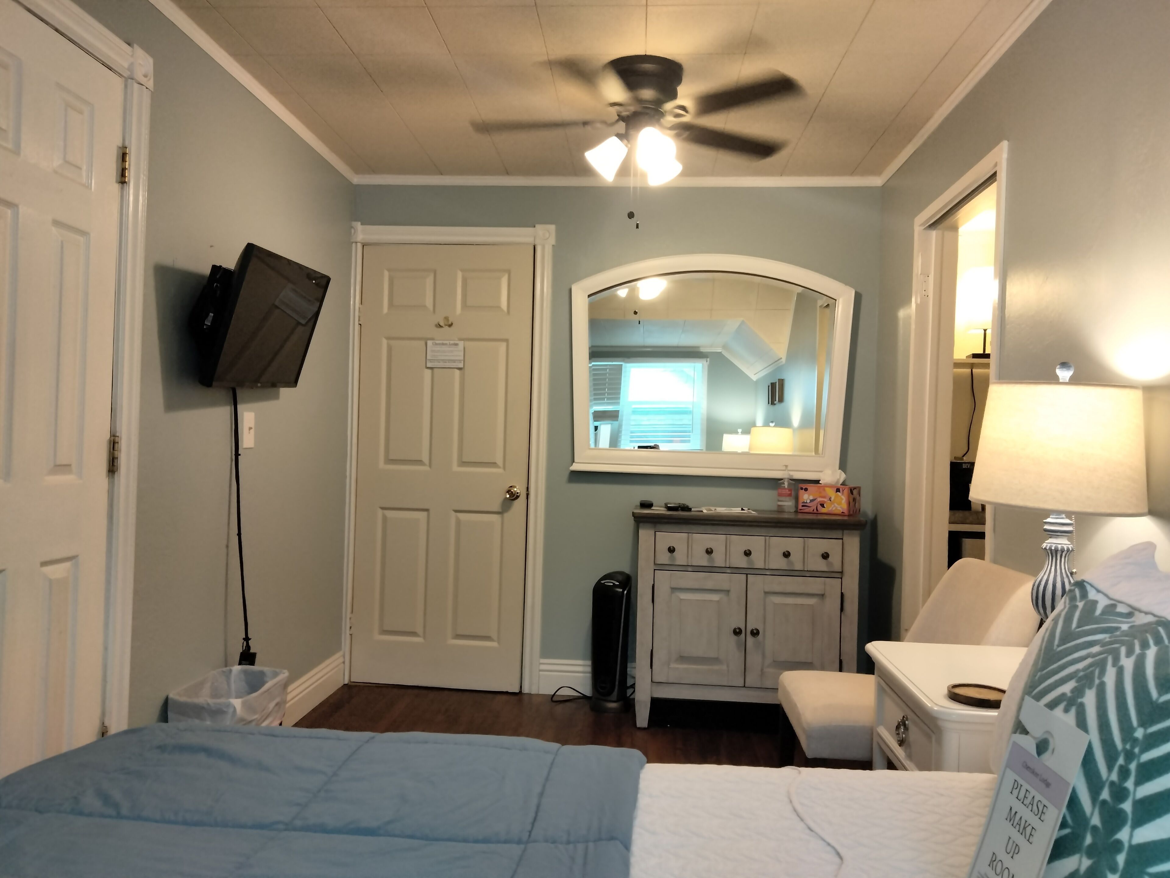 Un dormitorio con una cama, ventilador de techo, televisor montado en la pared, cómoda, silla y mesita de noche con lámpara.