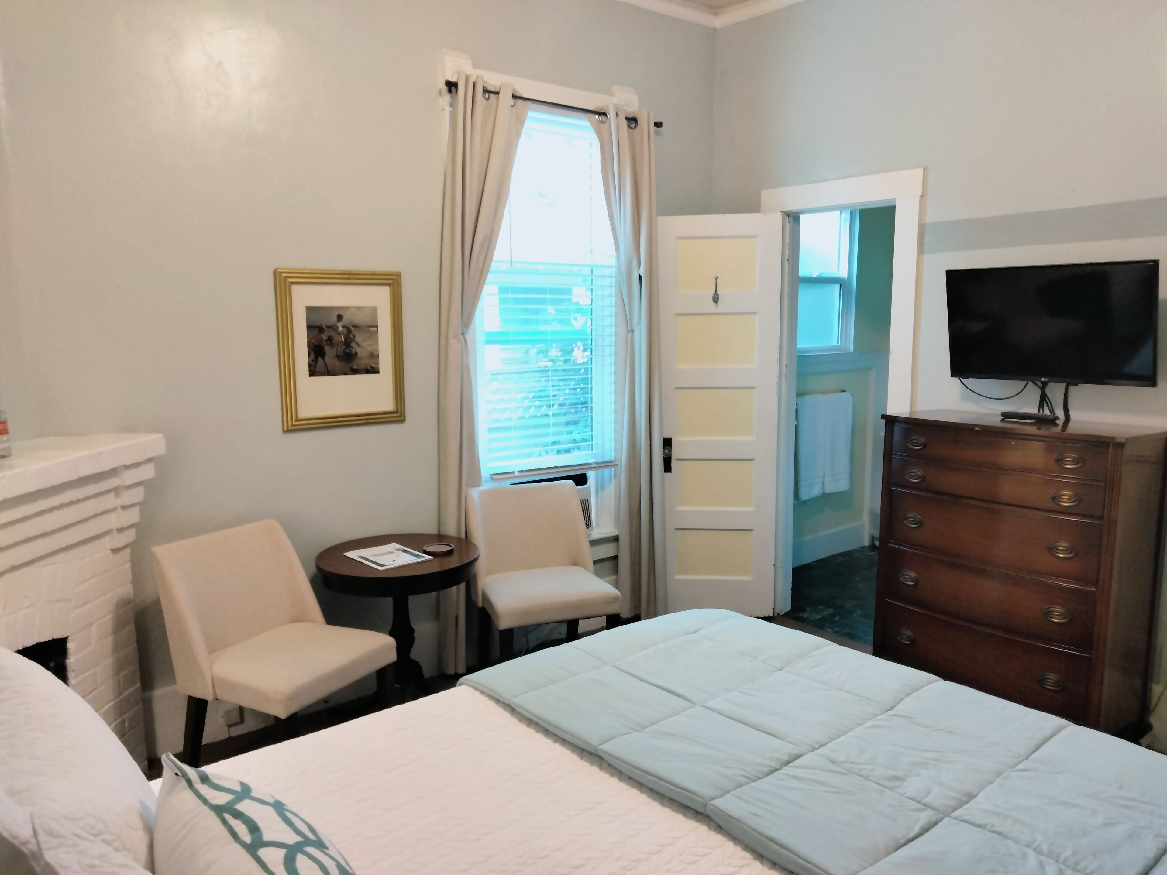 A hotel room with a bed, a chair, a small table, a dresser, a television, and a window with curtains.