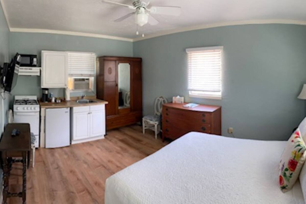 A bedroom with a bed, a dresser, a desk, a chair, a small kitchenette, and a ceiling fan.
