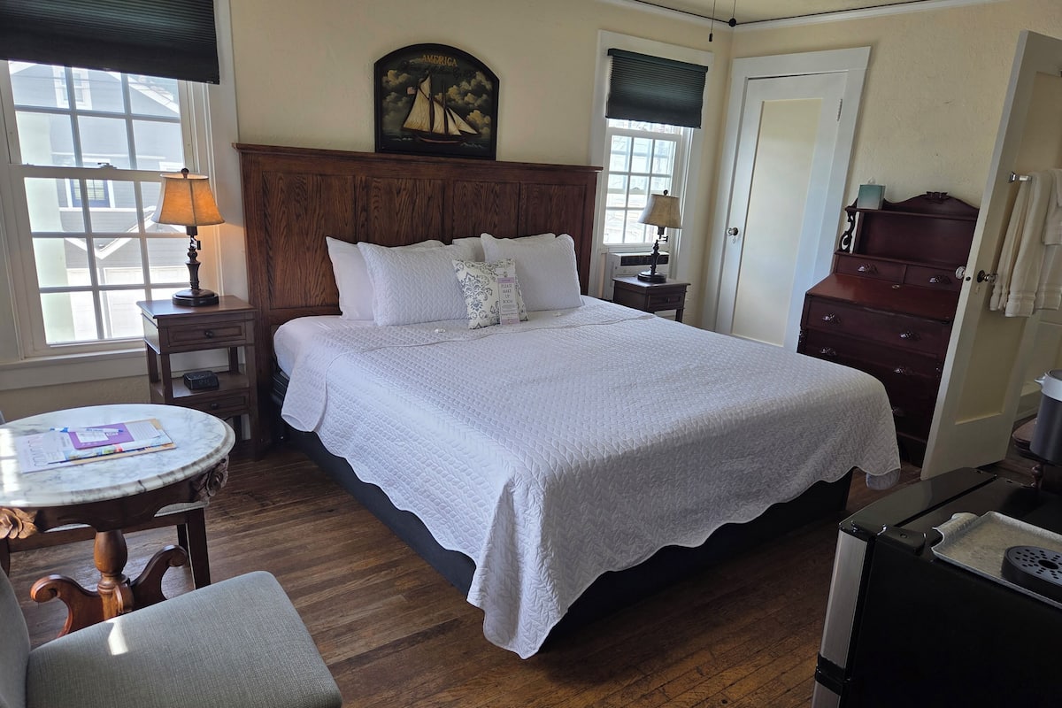 A neatly arranged bedroom with a large bed, a bedside table with a lamp, a small table with papers, and a chair.