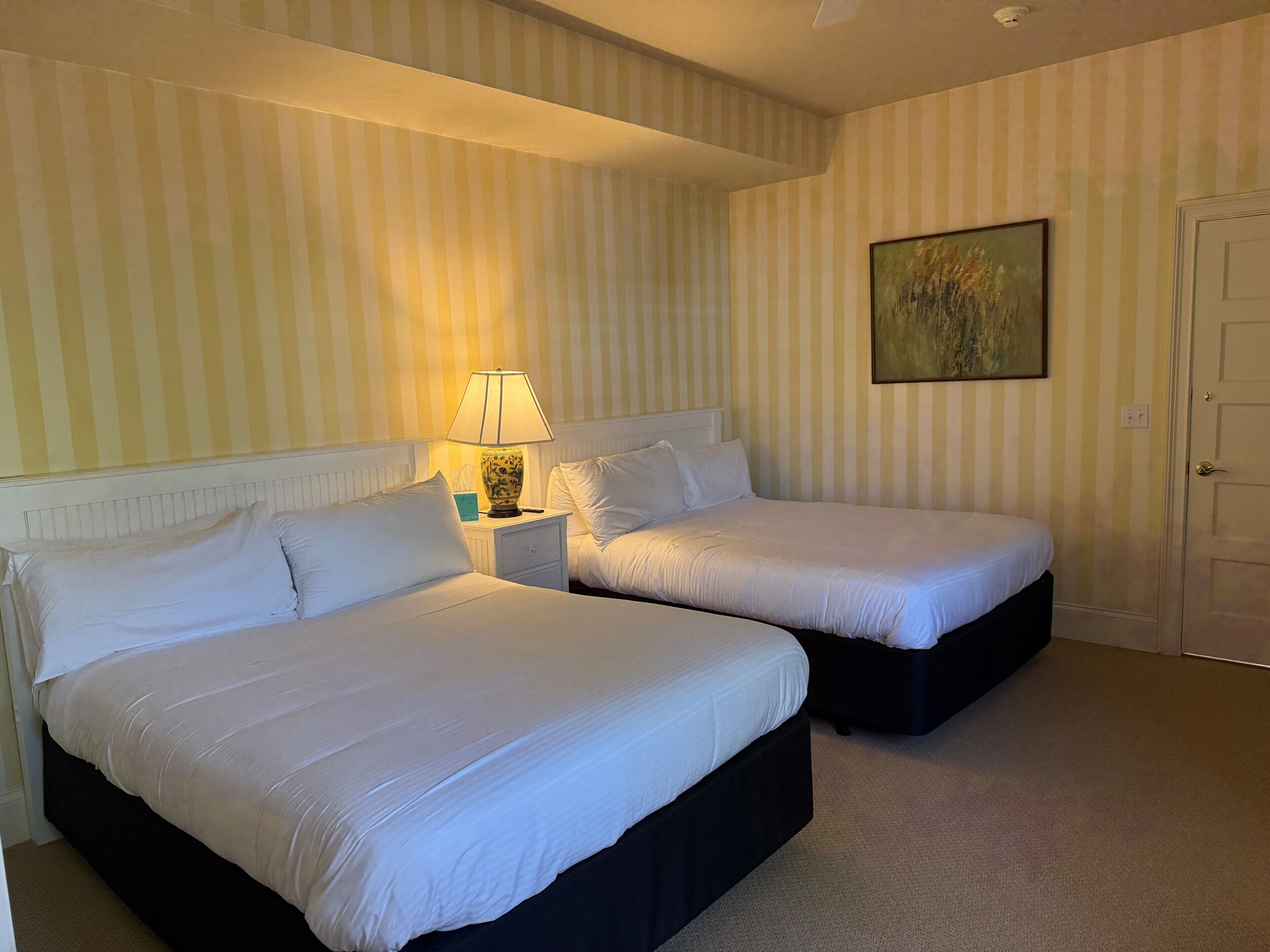 A hotel room with two beds, a nightstand with a lamp, a painting on the wall, and a door.