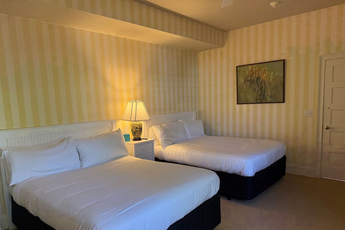 A hotel room with two beds, a nightstand with a lamp, a painting on the wall, and a door.