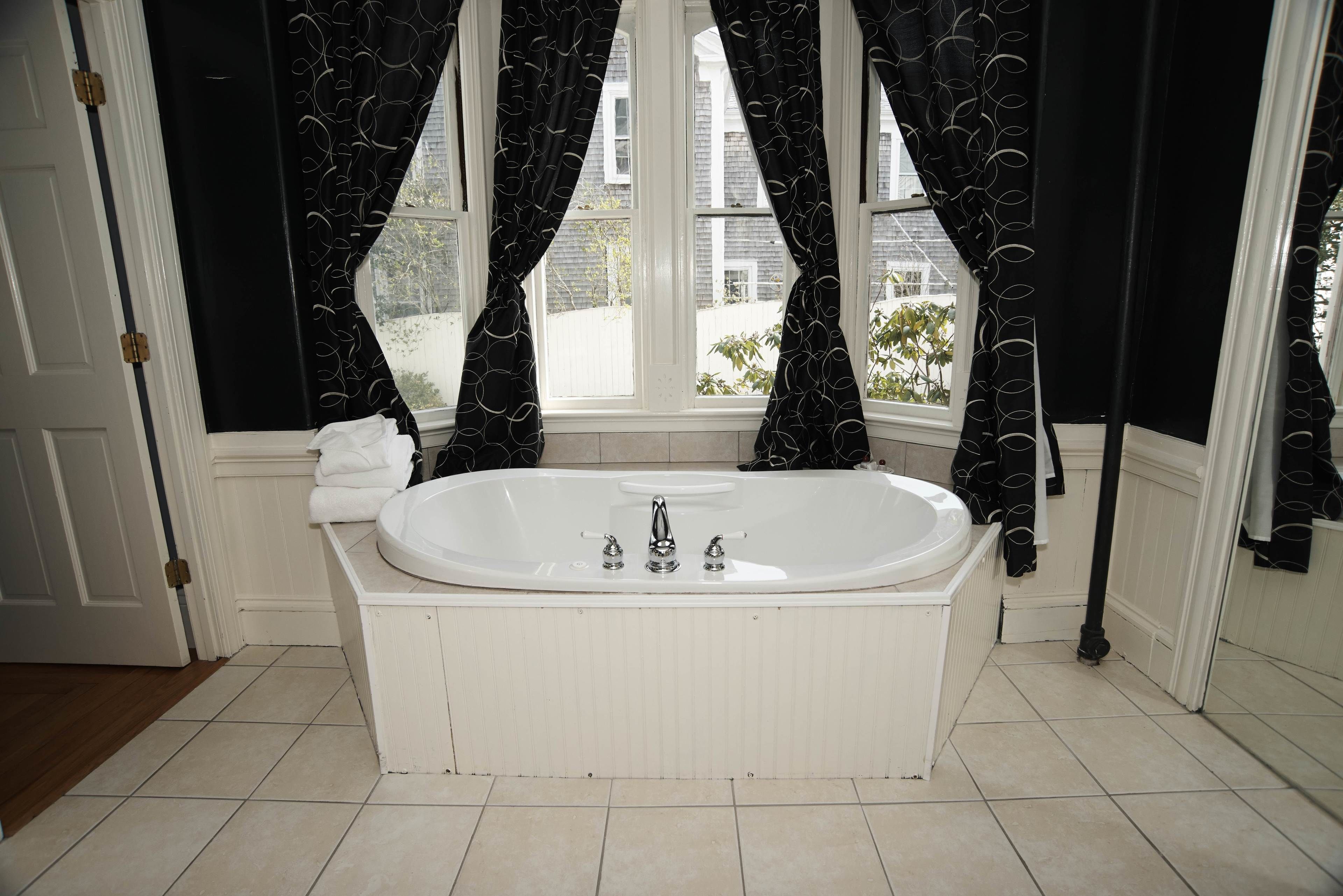 Ein Badezimmer mit freistehender Badewanne, weiß gefliestem Boden und dunkel gemusterten Vorhängen.