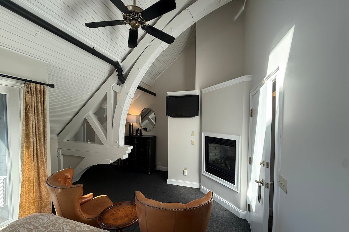 A bedroom with a bed, two chairs, a ceiling fan, a fireplace, and a TV.