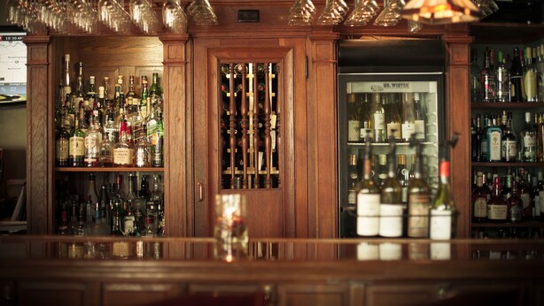 A bar area with shelves of liquor bottles, a wine cooler, and hanging wine glasses. - Belfry Inn & Bistro (Sandwich)