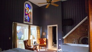 A hotel room with a large bed, a sitting area with a chair and table, a bathroom with a bathtub, a stained glass window, and a staircase leading to a lower level. - Belfry Inn & Bistro (Sandwich)