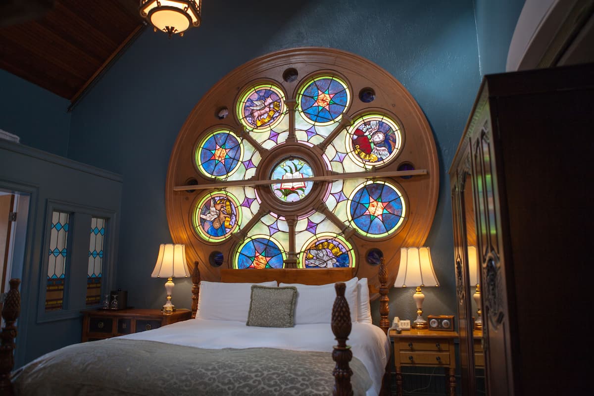 A bedroom with a large stained glass window above the bed, wooden headboard, and two bedside lamps.