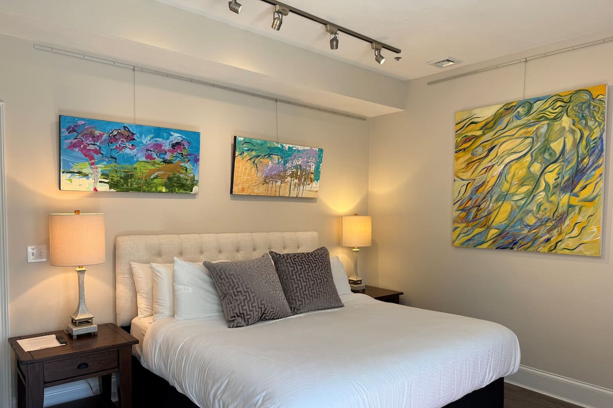 A bedroom with a large bed, two bedside lamps, a nightstand, and three paintings on the wall.