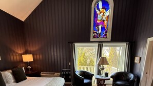 A bedroom with a large bed, two chairs, a small table, a ceiling fan, a stained glass window, and a view of the outdoors. - Belfry Inn & Bistro (Sandwich)