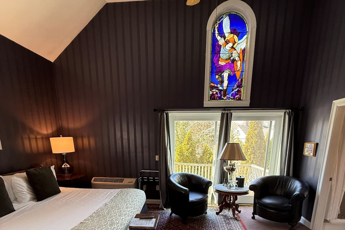 A bedroom with a large bed, two chairs, a small table, a ceiling fan, a stained glass window, and a view of the outdoors.