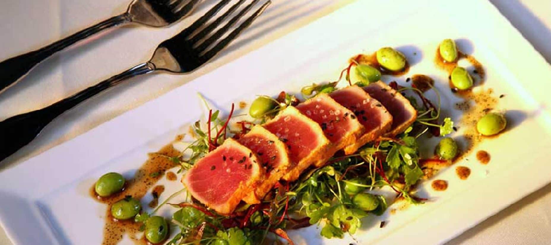 A white plate with a dish of sliced meat, greens, and olives.