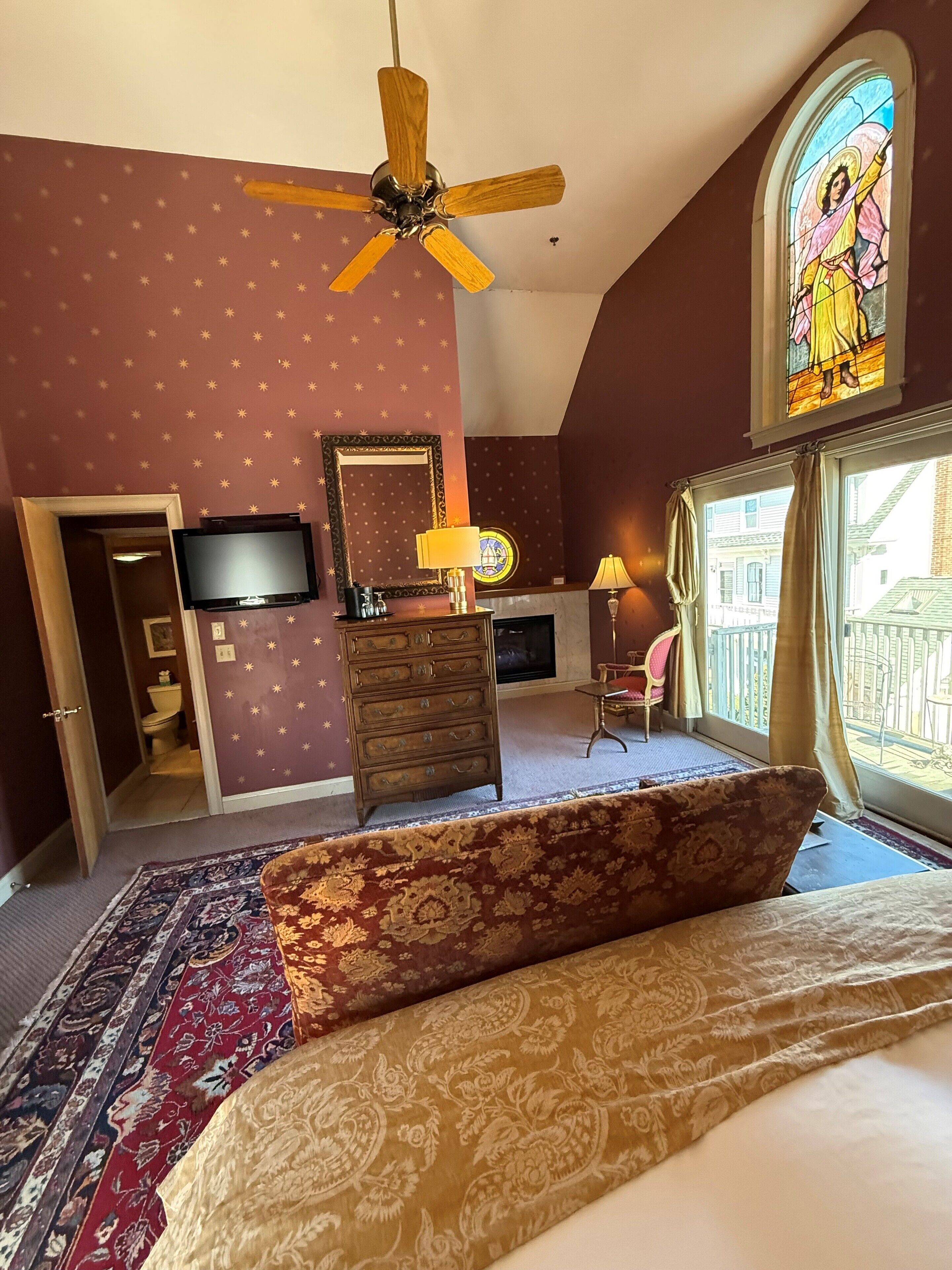A bedroom with a bed, a dresser, a television, a ceiling fan, a stained glass window, and a view of the outside.