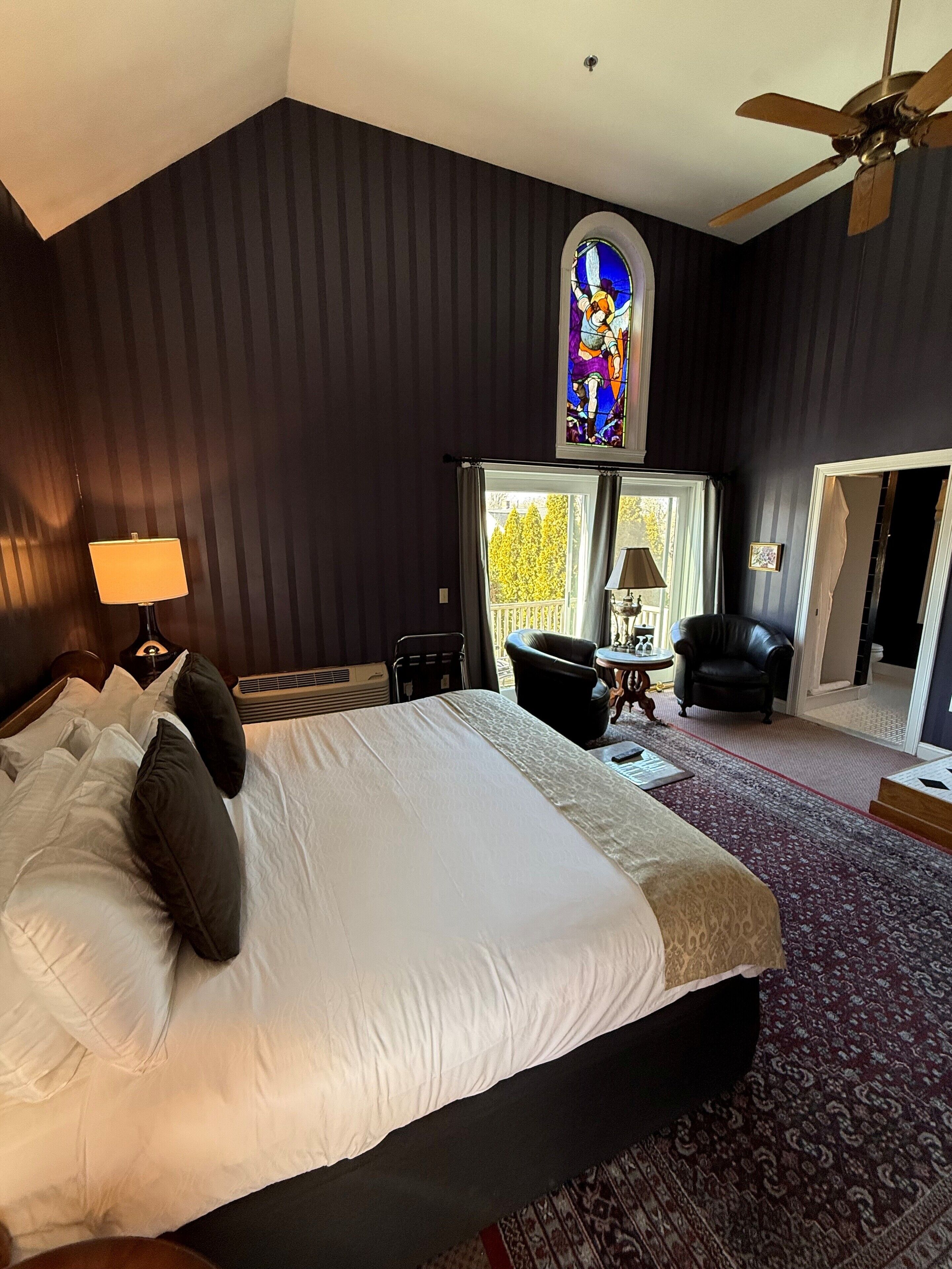 A hotel room with a large bed, a ceiling fan, a stained glass window, and a view of trees through the window.