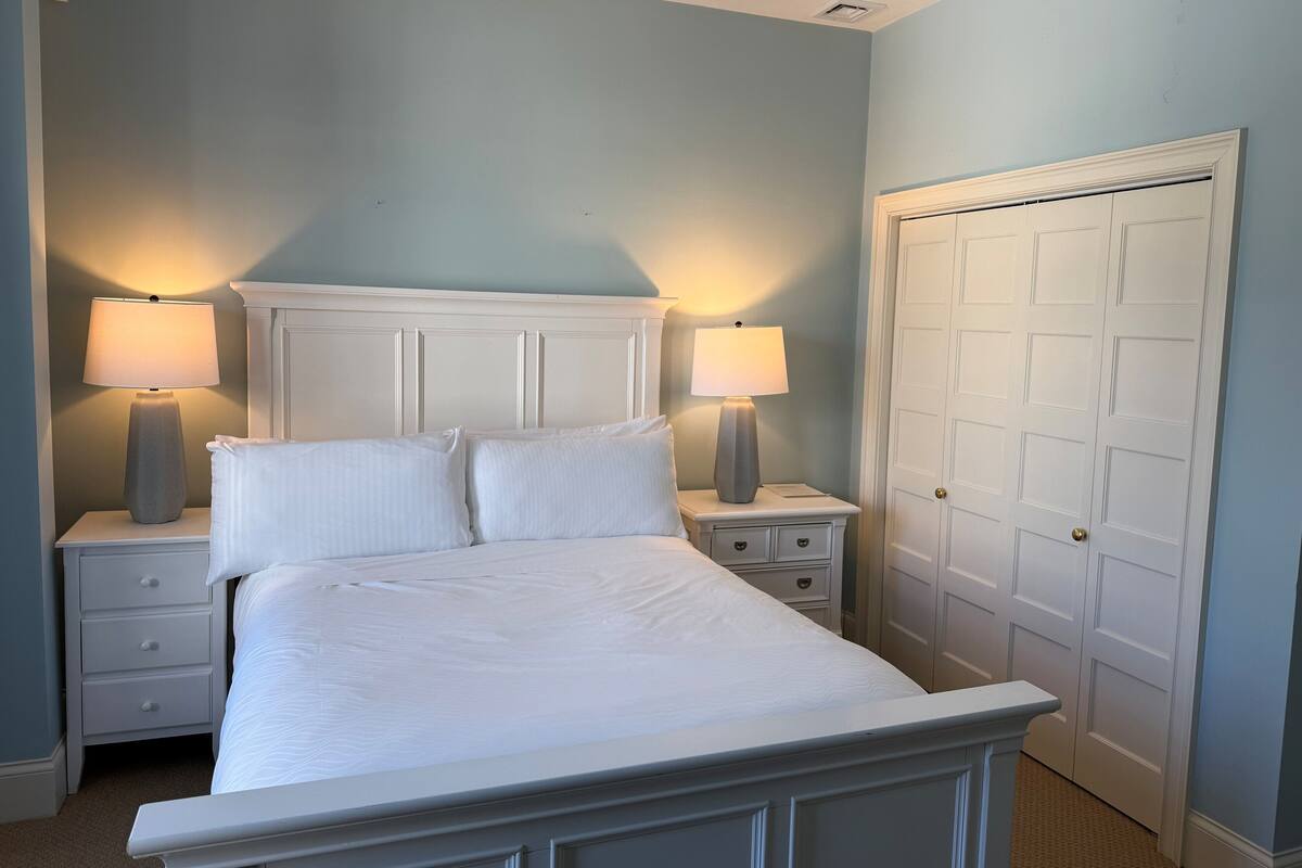 A bedroom with a bed, two bedside tables with lamps, a white door, and a light blue wall.