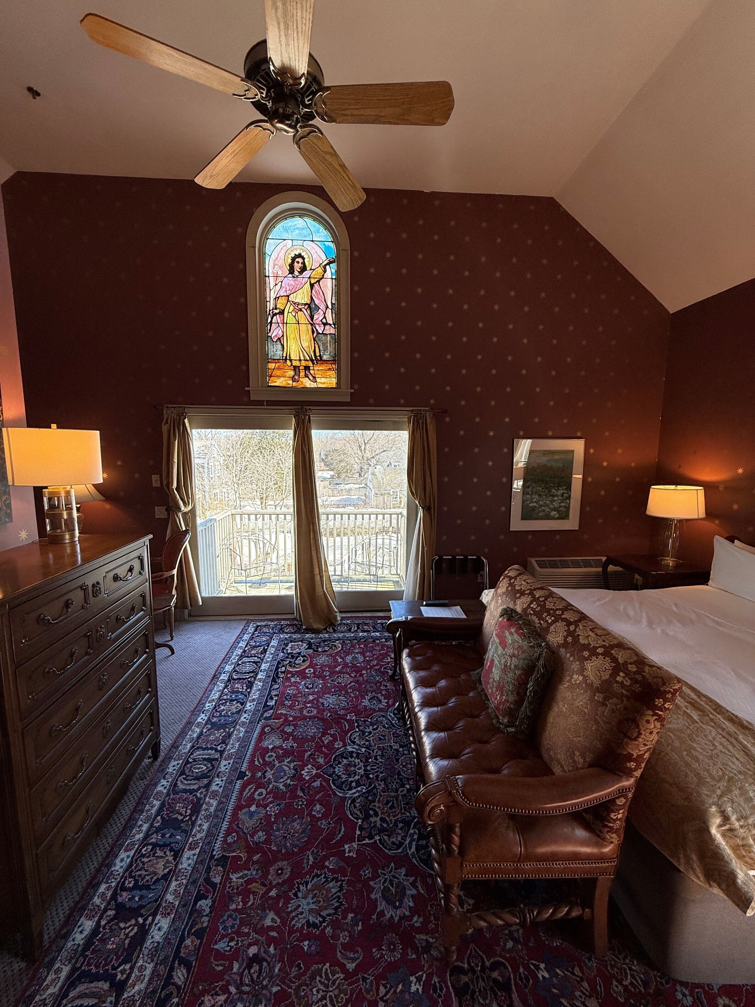 Ein Hotelzimmer mit einem großen Bett, einer Ledersofa, einem Holzkasten, einem Deckenventilator, einem Buntglasfenster und Blick nach außen.