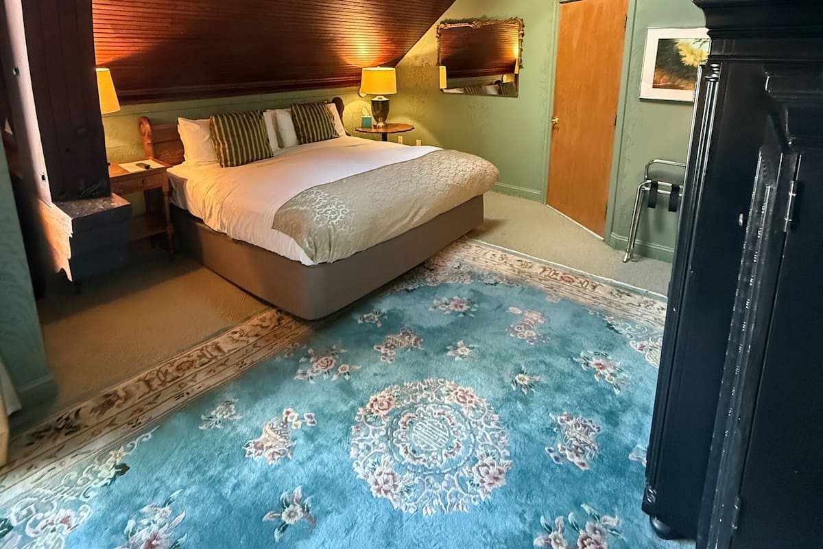 A bedroom with a sloped ceiling, a bed with a floral-patterned rug, a nightstand, a lamp, and a dresser.