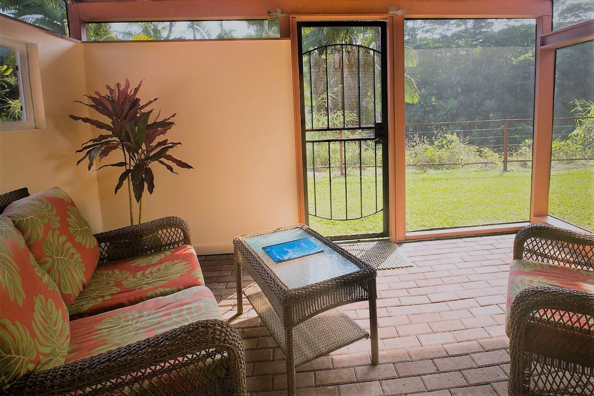 Un patio techado con muebles de mimbre, una mesa con un libro y vista a un jardín a través de una reja metálica.