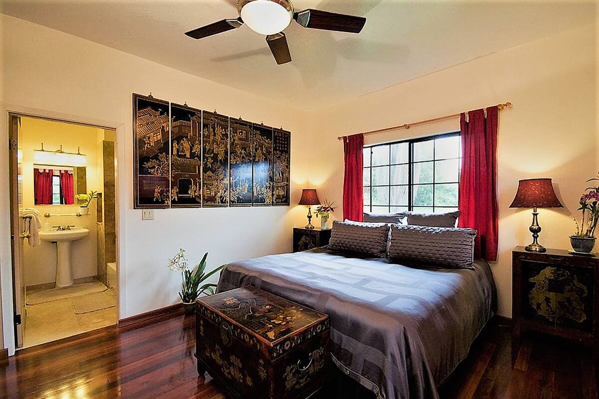 Un dormitorio con cama, ventilador de techo, ventana con cortinas rojas, mesita de noche con lámpara y un panel decorativo en la pared.