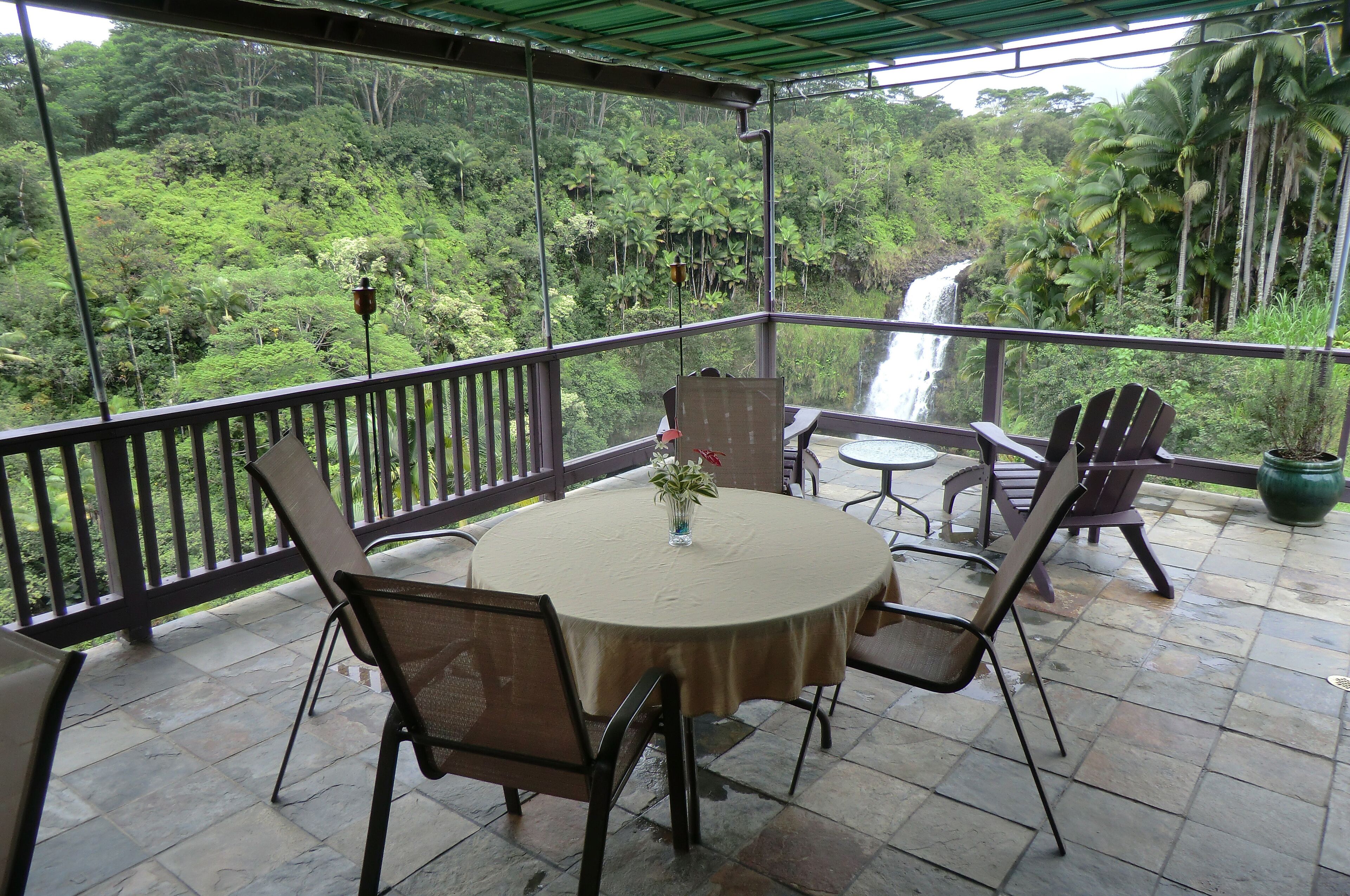 A covered outdoor seating area with a table and chairs, overlooking a lush forest and a waterfall.