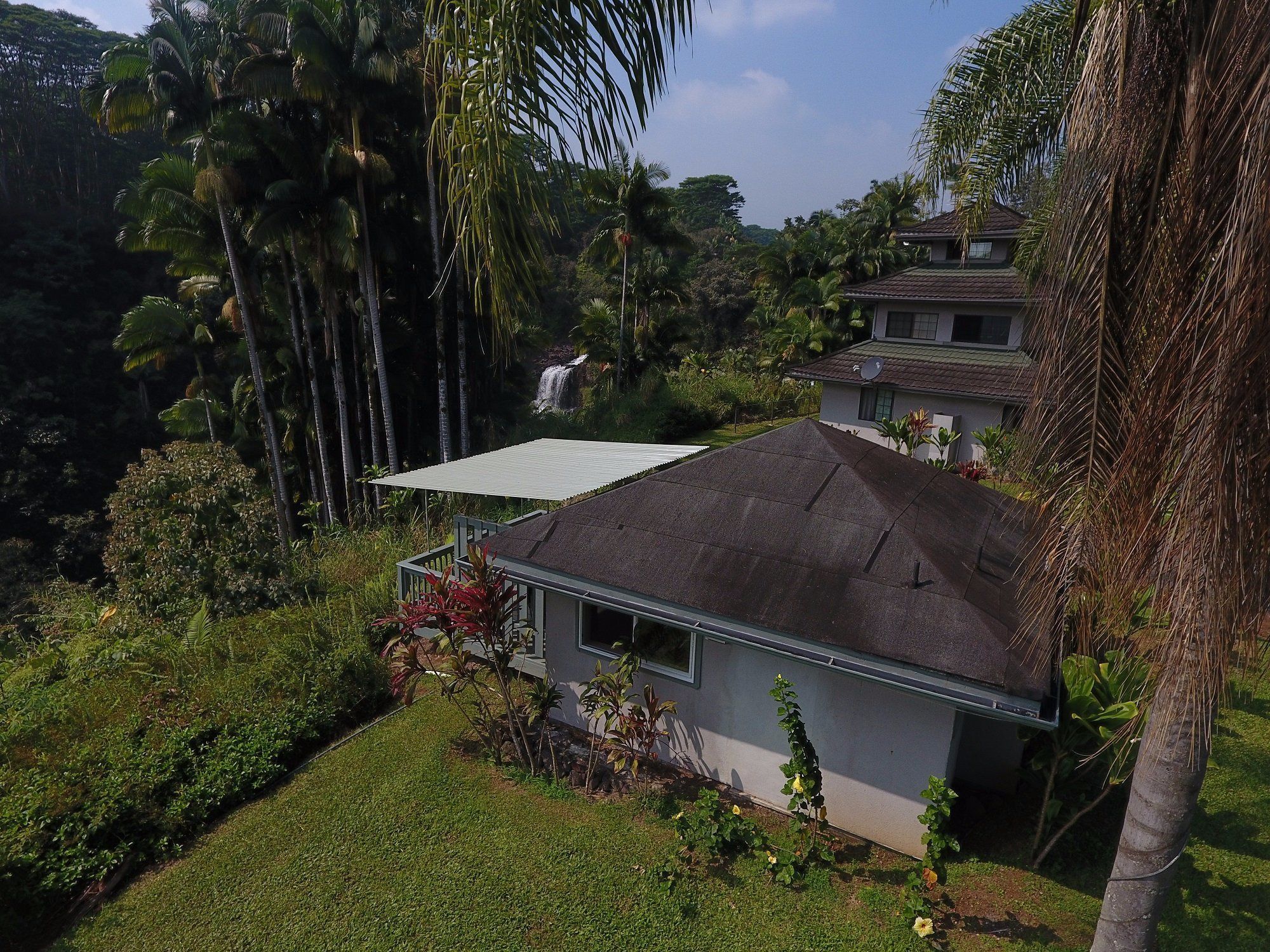 A house with a dark roof, surrounded by lush greenery and a waterfall in the background.