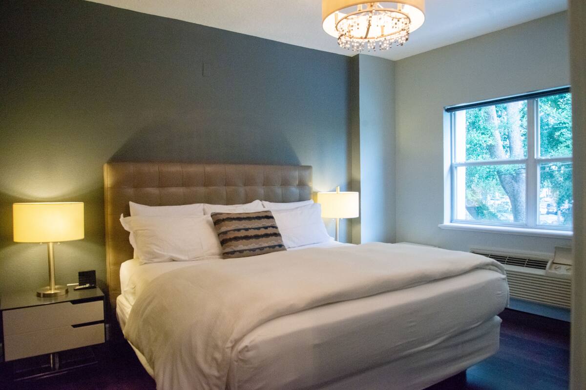 Un grand lit avec du linge de lit blanc, une tête de lit et une table de chevet avec une lampe.