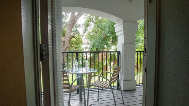 Une terrasse couverte, dotée d’une table et de chaises, entourée d’une balustrade blanche et offrant une vue sur la verdure.