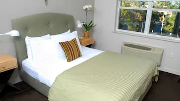 A neatly made bed with a green headboard, white bedding, and a striped blanket. There is a nightstand with a lamp and a small potted plant. A window with a view of trees and a building is visible.