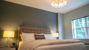 Une chambre d’hôtel avec un grand lit, deux lampes de chevet, un luminaire au plafond et une fenêtre donnant sur des arbres.