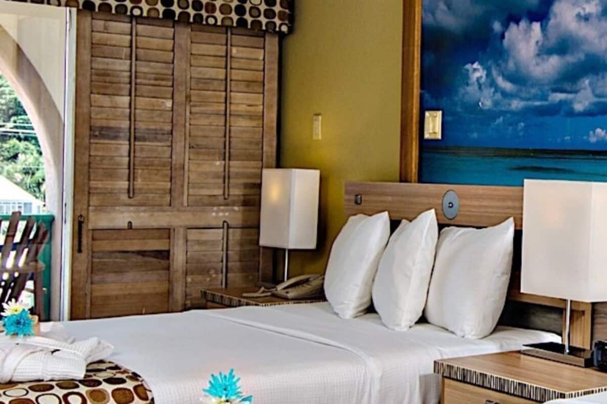 Ein Hotelzimmer mit Bett, Nachttischen, einer Lampe und einem großen Gemälde mit Strandblick an der Wand.
