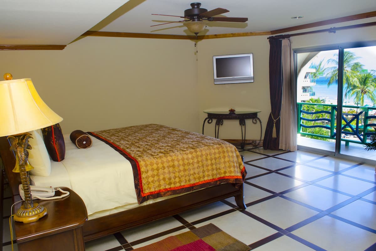 Ein Hotelzimmer mit Bett, Schreibtisch mit Lampe, Fernseher und einem Balkon mit Blick auf Palmen und Meer.