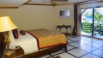 A hotel room with a bed, a desk with a lamp, a television, and a balcony with a view of palm trees and the ocean.