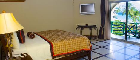 Habitación de hotel con cama, escritorio con lámpara, televisión y balcón con vista a palmeras y al océano.