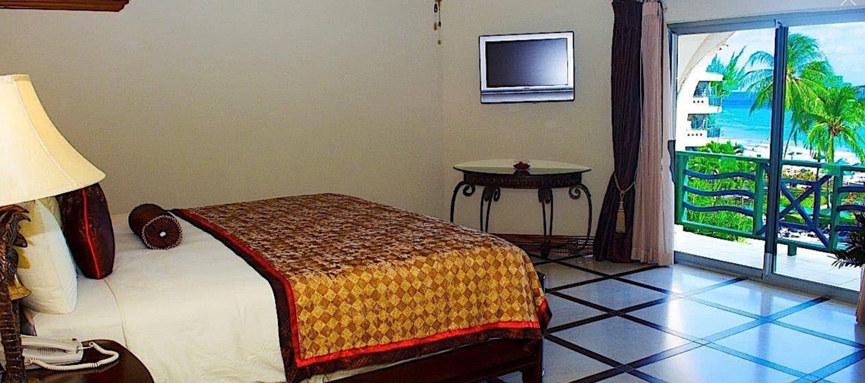 A hotel room with a bed, a bedside lamp, a television, a small table, and a view of a beach through a large window.